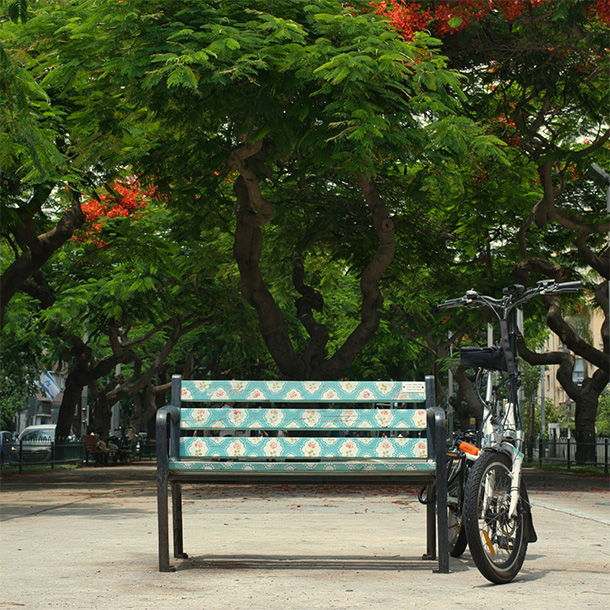 An electric bicycle tied to a bench, its planks covered with teal wallpaper and flower patterns, on concrete ground, under green trees in a boulevard