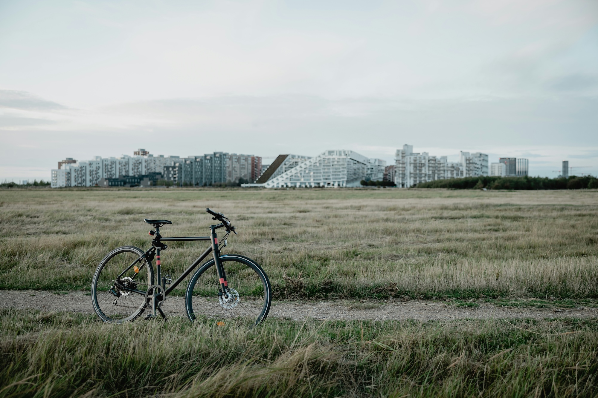 Overcast skies over a grassy field, modern city buildings in the background; a bicycle stands, propped on its kickstand, in the middle of a gravel path.