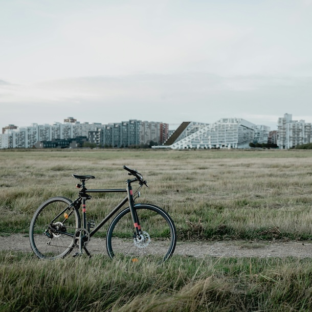 Overcast skies over a grassy field, modern city buildings in the background; a bicycle stands, propped on its kickstand, in the middle of a gravel path.