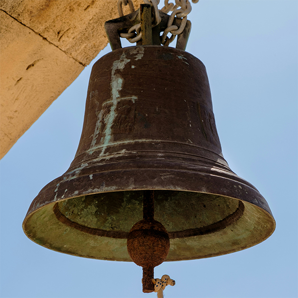In front of a clear blue sky, off the side of a building made of beige stone, a large bell with streaks of green patina hangs off a chain